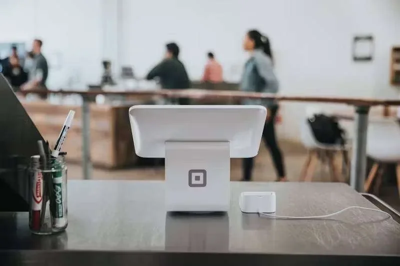 Terminal de pagamento branco em balcão metálico, com canetas em pote e pessoas desfocadas ao fundo
